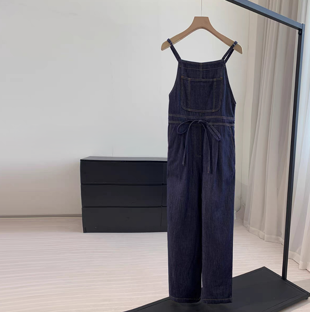 A dark blue denim jumpsuit with adjustable shoulder straps and a matching waist tie, hanging on a wooden hanger on a clothing rack in a minimalist room.